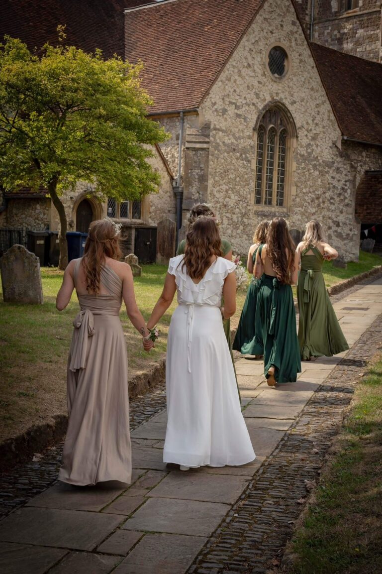 A bride and bridesmaids in sage and green dresses walking along a churchyard path, Beatrice Espinosa Bridal