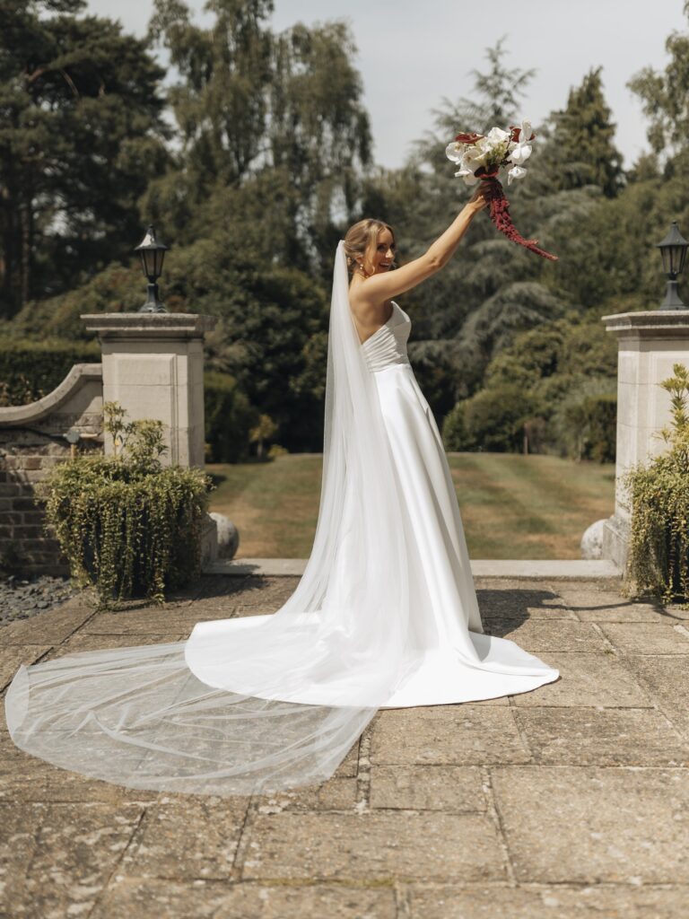 A bride with a long veil and train raising her bouquet in a formal garden with stone gate posts, Beatrice Espinosa Bridal