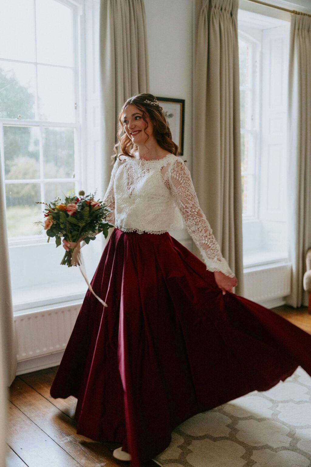 A bride in a lace top and a full burgundy silk skirt standing in a bright window