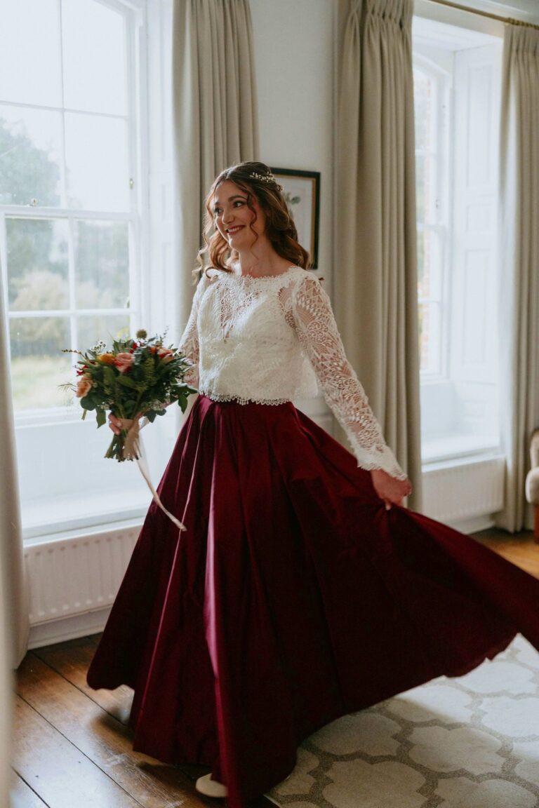 A bride in a lace top and a full burgundy silk skirt standing in a bright window, Beatrice Espinosa Bridal