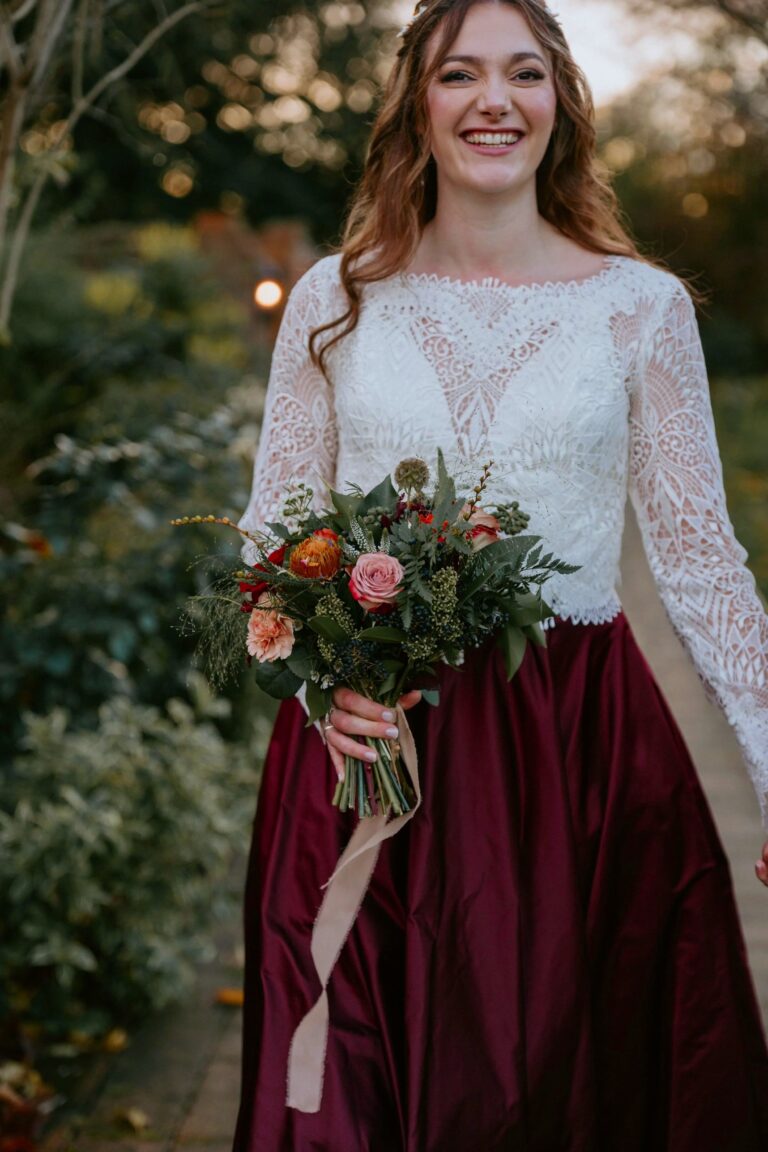 A bride in a lace-sleeved top and a burgundy skirt holding an autumn bouquet outdoors, Beatrice Espinosa Bridal