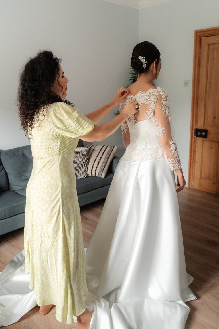 A bride on the morning of her wedding having the lace-sleeved gown fastened by her dressmaker, Beatrice Espinosa Bridal