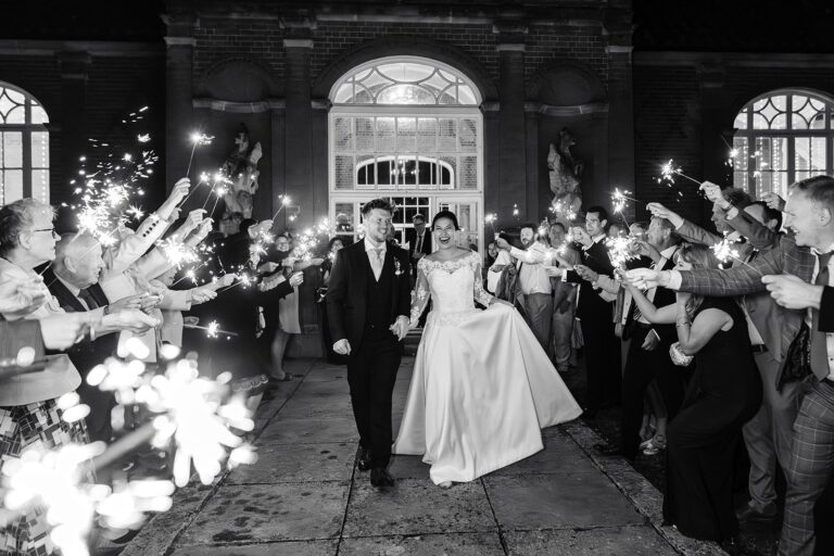 A couple in black and white walking between guests holding sparklers, the bride in a full silk gown, Beatrice Espinosa Bridal