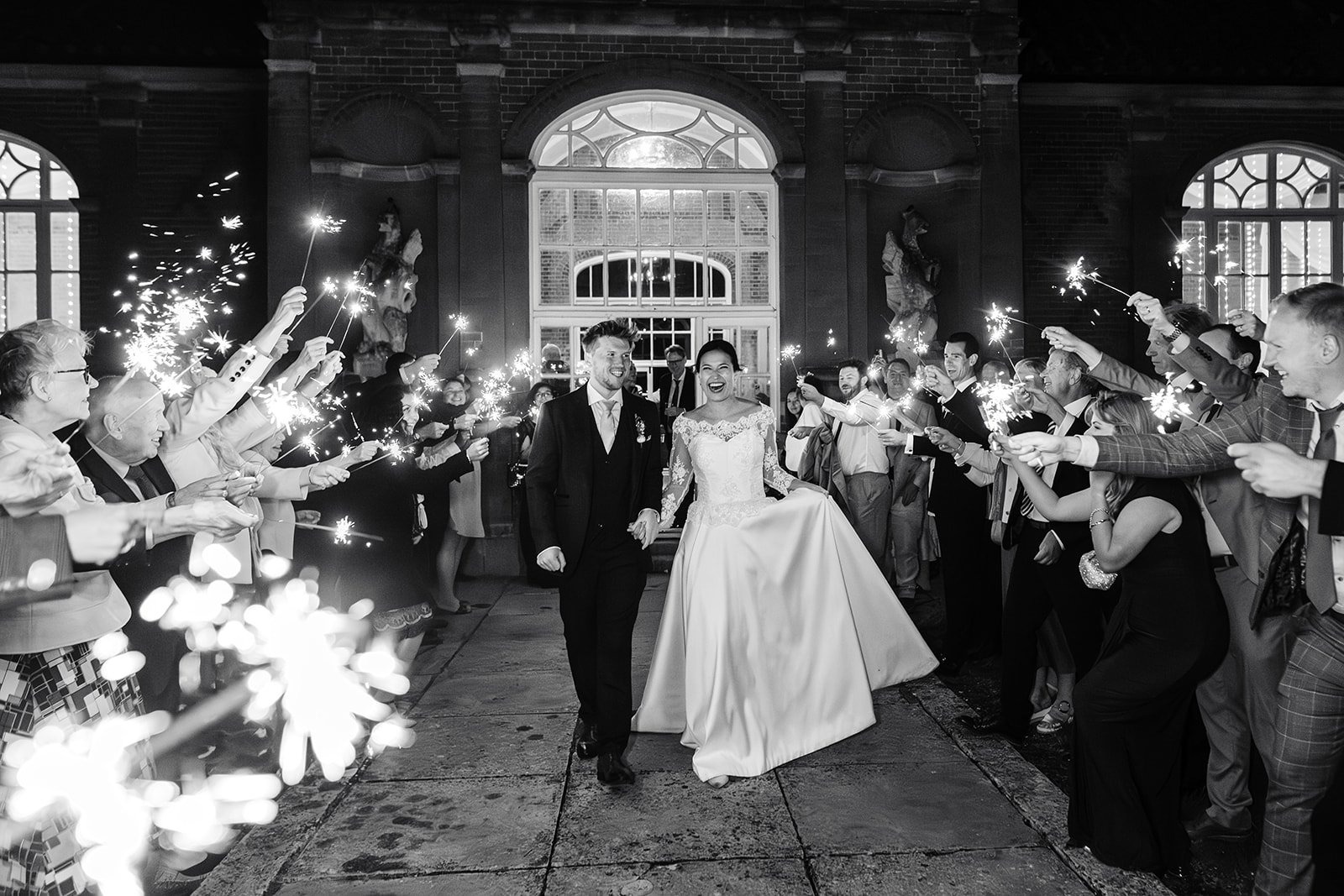 A couple in black and white walking between guests holding sparklers, the bride in a full silk gown, Beatrice Espinosa Bridal