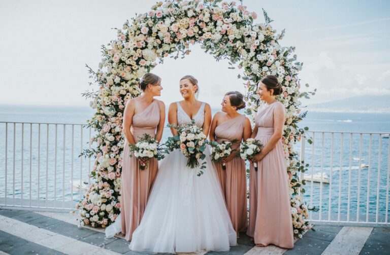 The bride and her bridesmaids under a rose arch, the sea and moored boats behind them, Bella Sposa