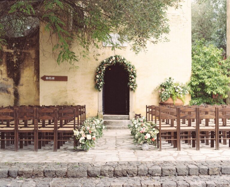 Ceremony chairs set before a floral arch in a stone courtyard at Danilia Village, Belli Momenti