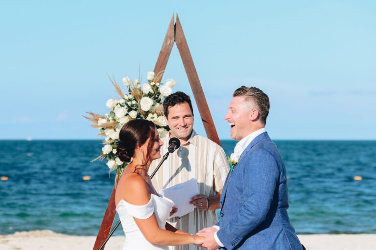 A couple and their officiant mid-ceremony on a beach with a wooden triangle arch, Better Together Mexico