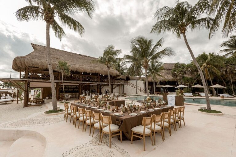 A long reception table beneath palm trees by a thatched palapa, Better Together Mexico