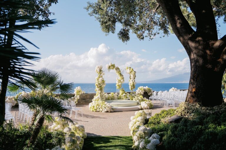 A ceremony platform under a white floral arch above a bay, Bianco Bouquet Weddings and Events