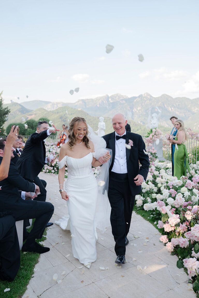 The bride on her father's arm through a shower of petals, mountains behind, Bianco Bouquet Weddings and Events
