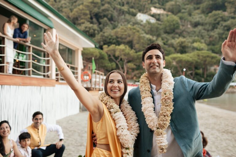 The couple in garlands raising their arms, guests on the sand, Bianco Bouquet Weddings and Events