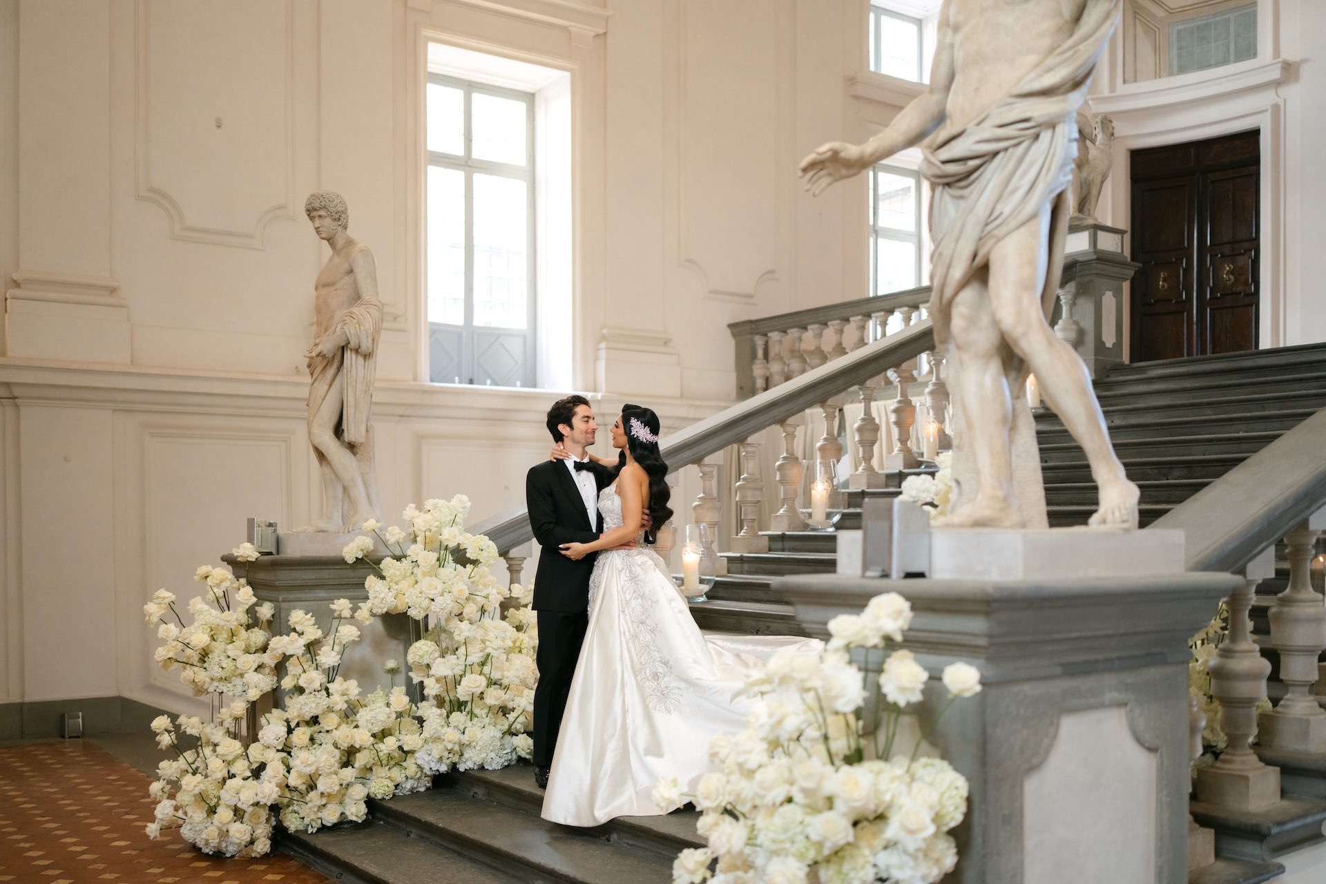The couple on a stone staircase between marble statues, white roses banked on the steps, Bianco Bouquet Weddings and Events