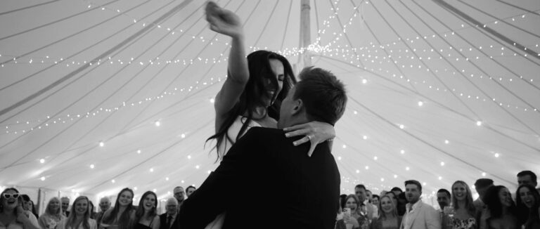 A first dance under the fairy lights of a marquee, guests gathered round, in black and white (frame from the film on their homepage), Blue Ridge Films