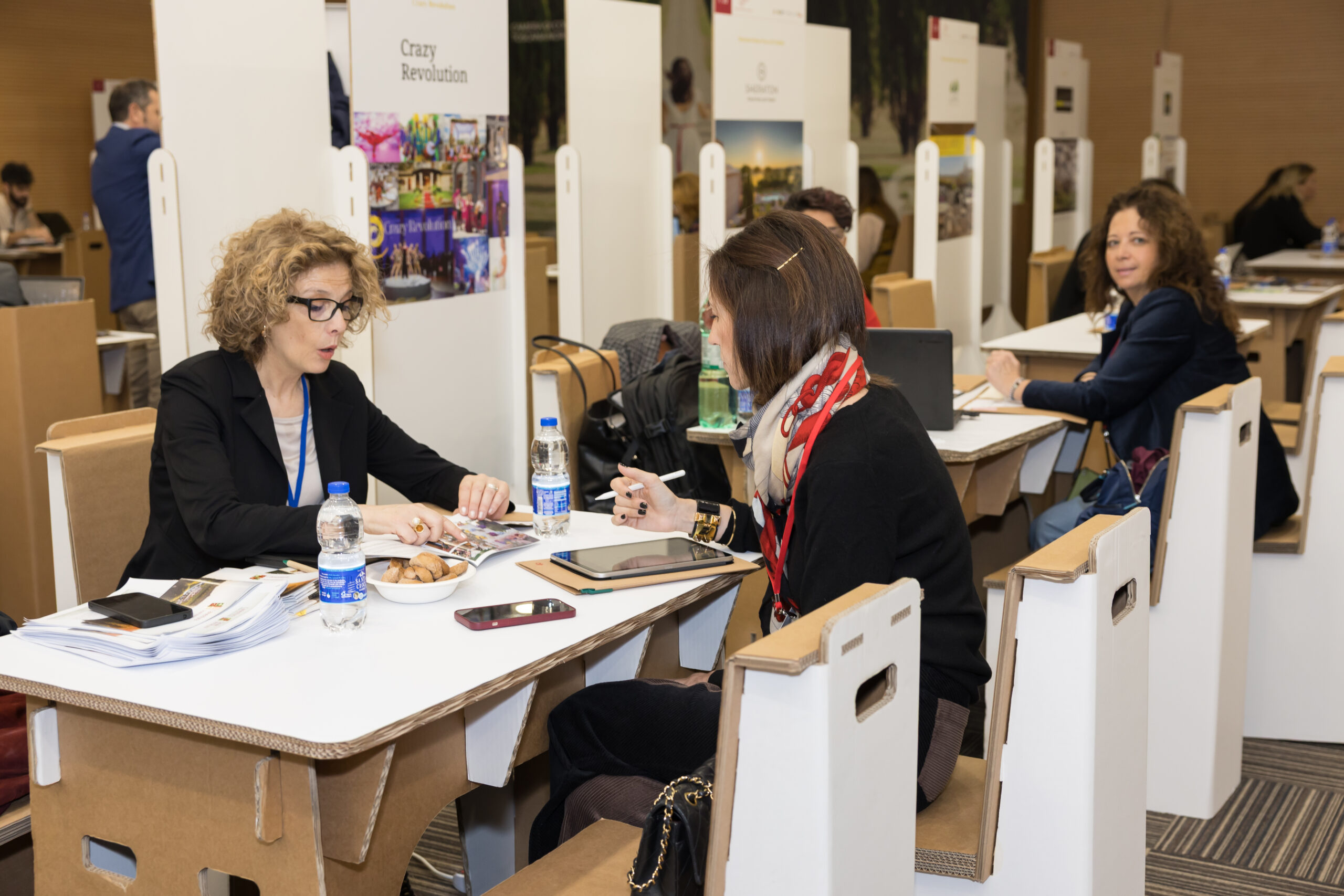 Two participants at a meeting table with a laptop and tablets, and other meetings behind them