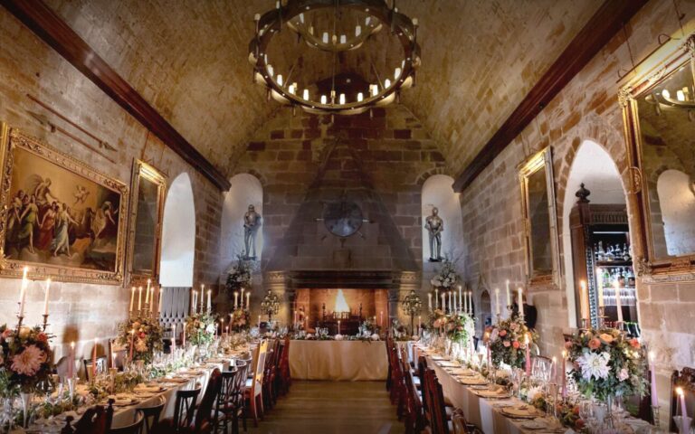 The Great Hall laid with long tables, candelabra and flowers beneath its pointed stone vault, with the fire lit at the far end, Borthwick Castle