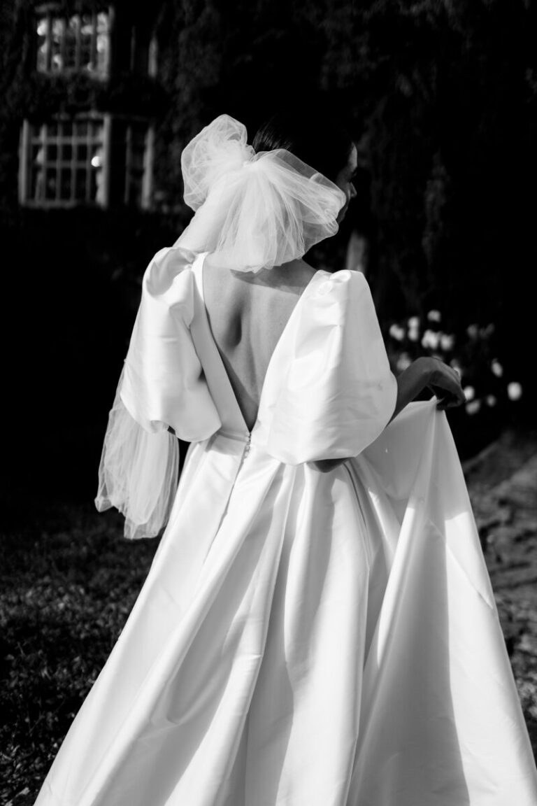 A veil catches the evening light, back turned to a stone facade, Botias
