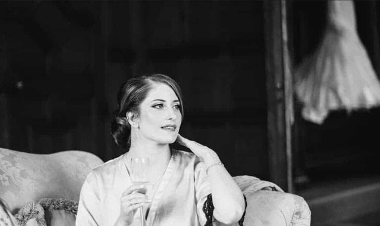 A bride rests in a wing chair before an antique mirror, robe loosely tied, Botias