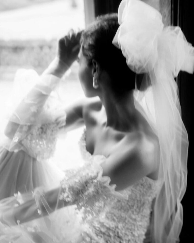 A hand raised to a bow-topped veil, caught at a window, Botias
