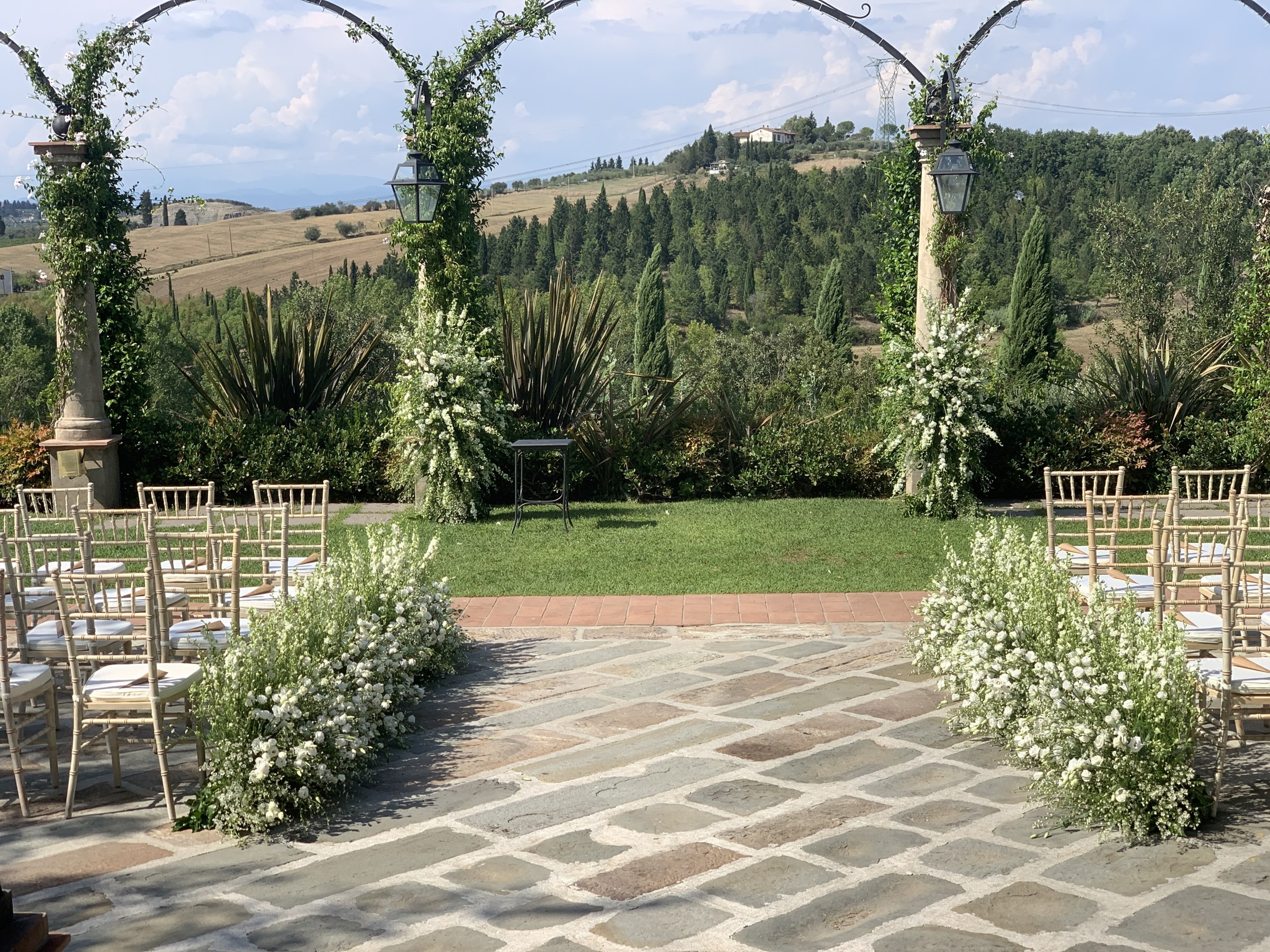 Ceremony aisle lined with white flowers beneath a pergola, with Tuscan hills beyond, Bottega dei Fiori by Ezio De Angelis