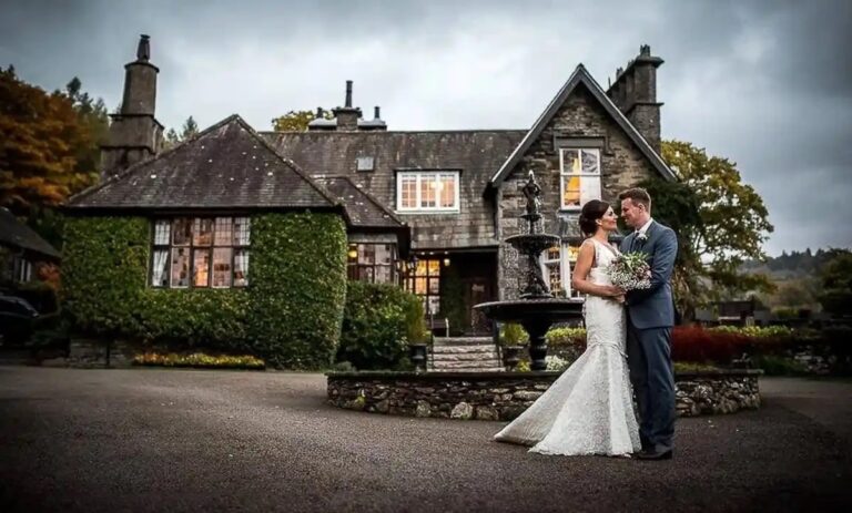 The couple before the ivy-clad stone house at dusk, the windows lit and the tiered fountain beside them, Broadoaks Country House