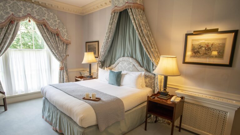 A Melbourne Lodge bedroom with a canopied double bed, swagged curtains and a sporting print on the wall, Brocket Hall