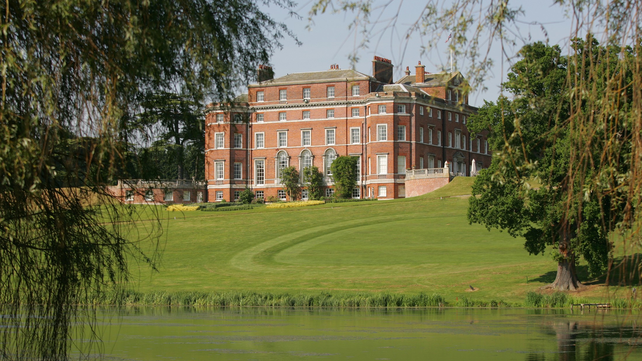 Brocket Hall on its rise above the Broadwater, seen across the water between willow and mature trees, Brocket Hall