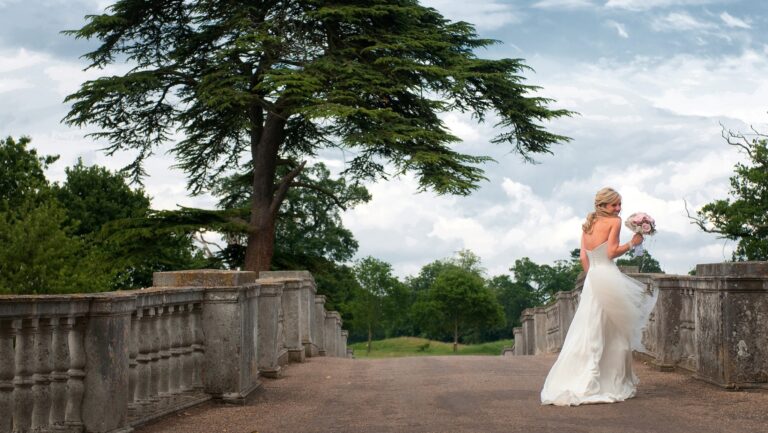 A bride holding her bouquet on a stone balustraded bridge in the park, under a spreading cedar, Brocket Hall
