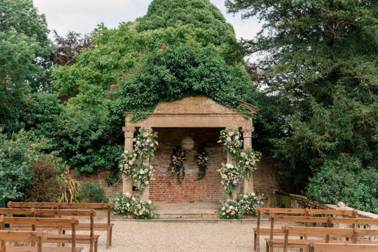 The Temple in the grounds, a stone portico against a brick back wall, garlanded with flowers and set with benches, Brympton House