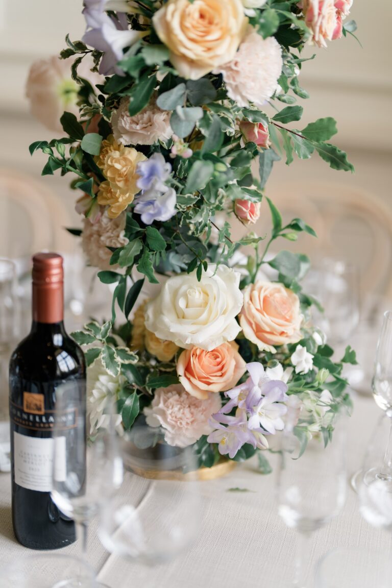A column of roses, sweet peas and foliage on a dining table, with a bottle of red wine beside it, Brympton House