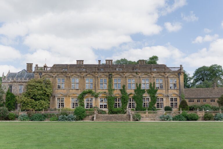The south front of Brympton House across the lawn, honey-coloured Ham stone with ivy between the tall windows and a balustraded terrace below, Brympton House