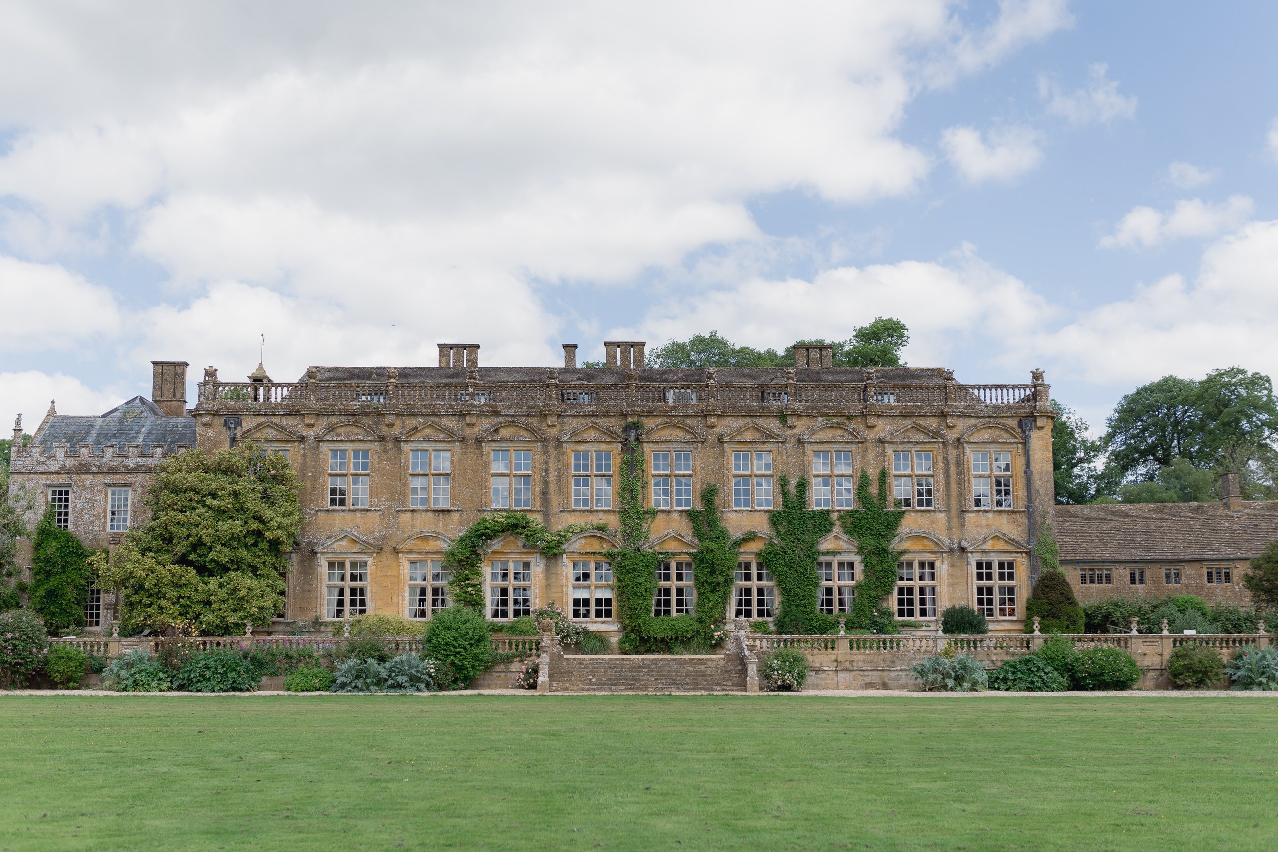 The south front of Brympton House across the lawn, honey-coloured Ham stone with ivy between the tall windows and a balustraded terrace below, Brympton House