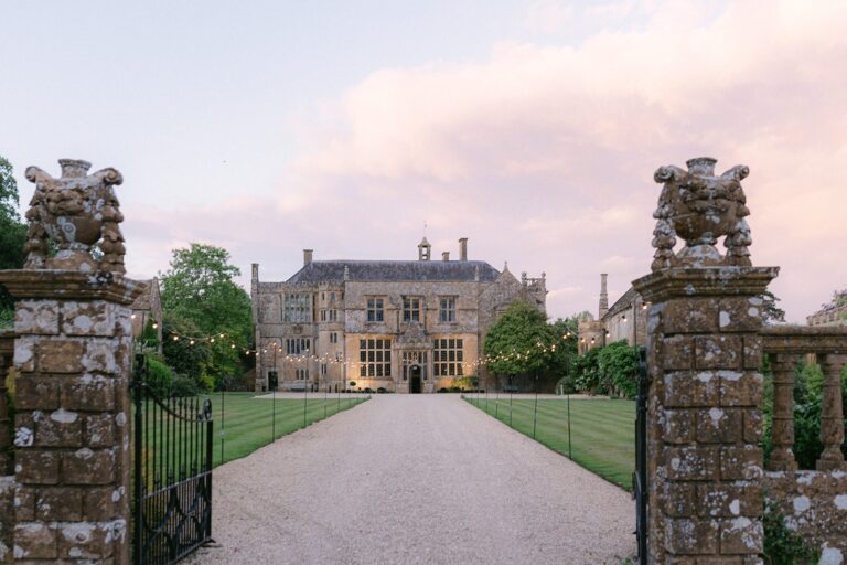 The drive to the house framed by carved stone gate piers, with festoon lights in the trees at dusk, Brympton House