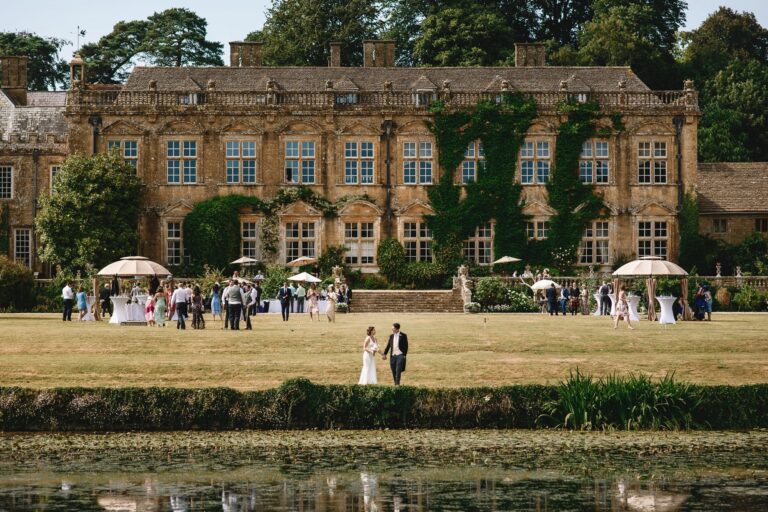 A couple on the grass beside the lake, with the south front and a drinks reception under white parasols behind, Brympton House