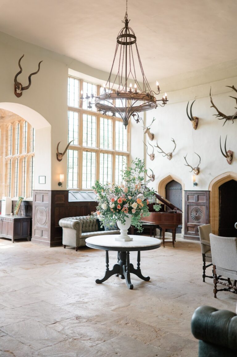 The Great Hall with its iron chandelier, flagstone floor, piano and antler trophies, set with a large flower arrangement, Brympton House
