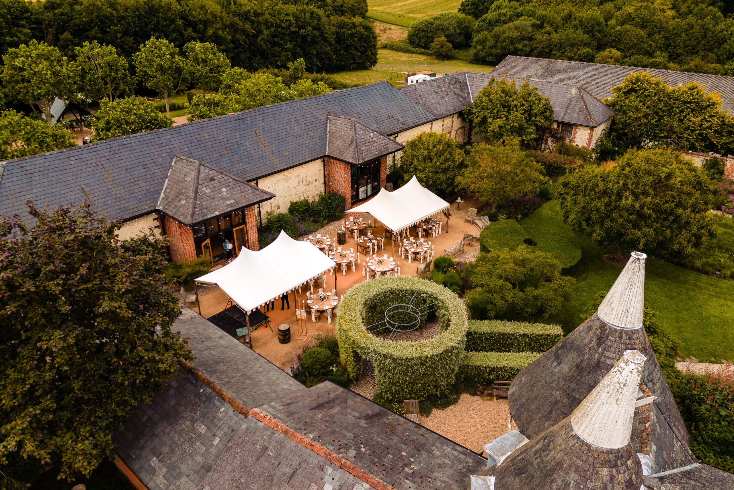 The courtyard from above, set with tables under two white stretch tents between the barn ranges and a round clipped hedge, Bury Court