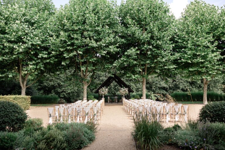 Outdoor ceremony set out on gravel beneath a row of plane trees, with an oak-framed arbour at the end of the aisle, Bury Court