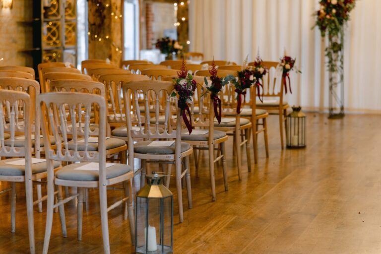 Chairs lined up either side of an aisle on the oak floor of the Barn, with lanterns and red flowers, Bury Court