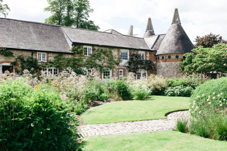 The stone and brick house with clipped box and a curving lawn, the oast house cones rising behind, Bury Court