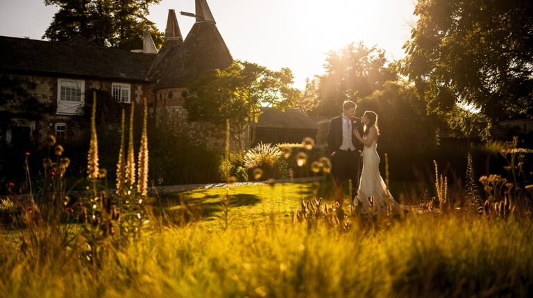 A couple standing in the long grasses of the garden at golden hour, the oast houses behind, Bury Court