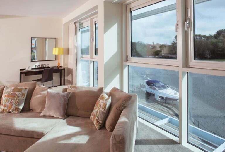A suite sitting area with a corner sofa and full-height glazing above a boat moored on the river, Captain's Club Hotel & Spa