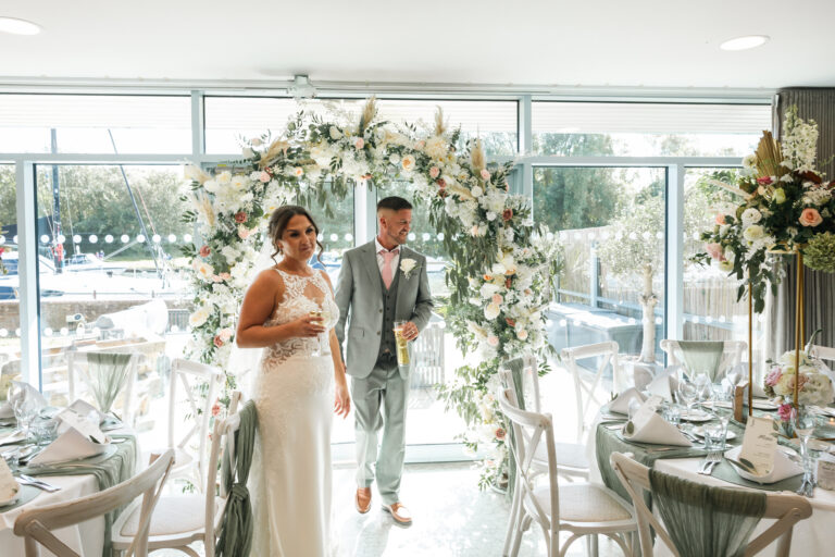 A newly married couple walking away from a floral arch at the window end of a function suite, with tables dressed in sage green, Captain's Club Hotel & Spa