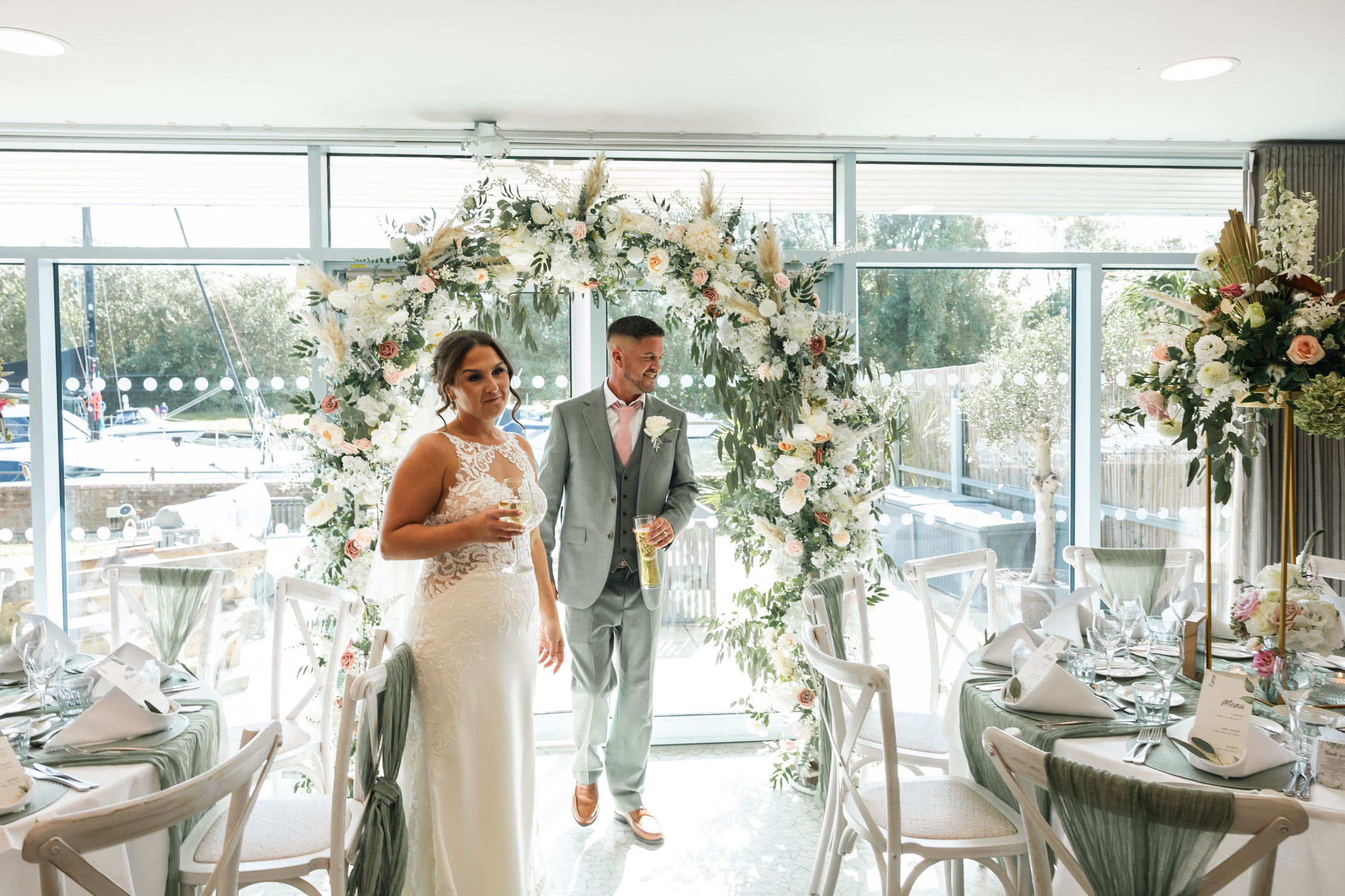 A newly married couple walking away from a floral arch at the window end of a function suite, with tables dressed in sage green, Captain's Club Hotel & Spa