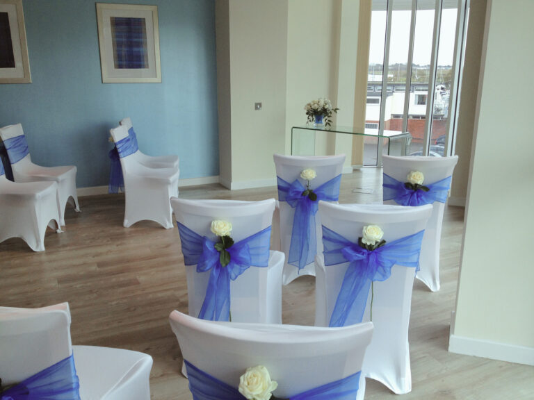 Rows of white-covered chairs tied with blue sashes and white roses, set for a small ceremony beside a river-view window, Captain's Club Hotel & Spa