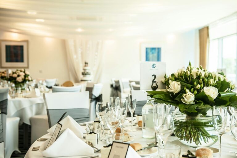 A wedding table centre of white roses in a glass bowl, with folded napkins, menu cards and a cake visible behind, Captain's Club Hotel & Spa