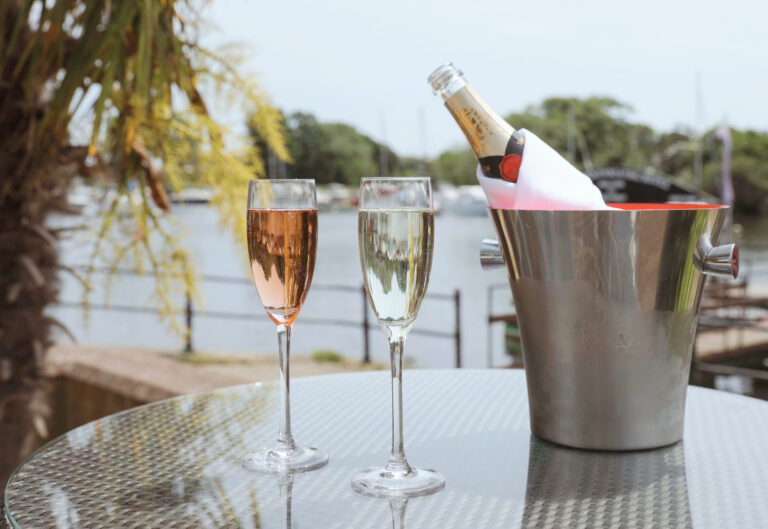 Two glasses of fizz and a bottle in an ice bucket on a terrace table, with the river and boats behind, Captain's Club Hotel & Spa