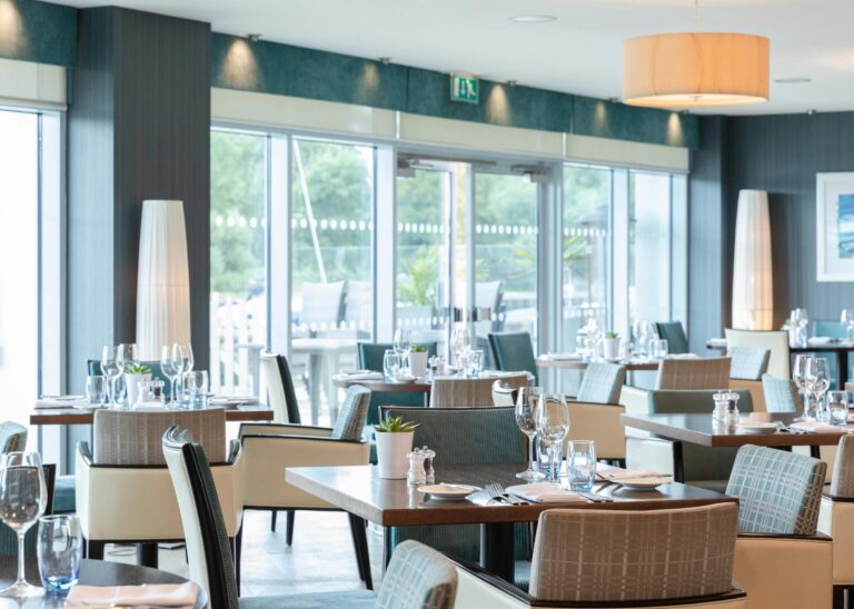 Tables laid in the restaurant beside full-height windows looking out to trees and the river, Captain's Club Hotel & Spa