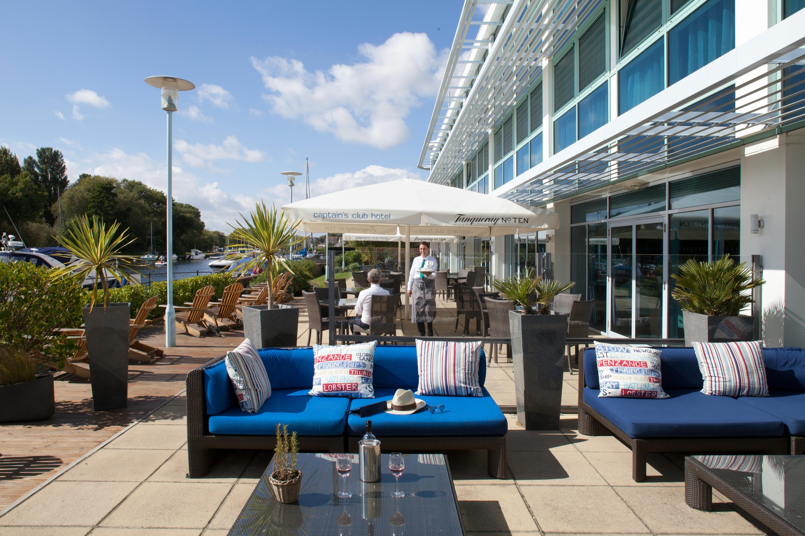 The riverside terrace with outdoor sofas and parasols, the hotel facade on the right and moored boats on the River Stour beyond, Captain's Club Hotel & Spa