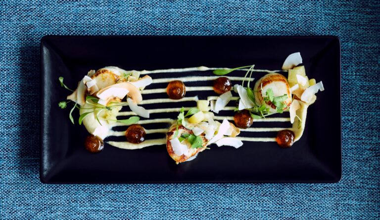 Seared scallops laid across a striped sauce on a dark rectangular plate, Captain's Club Hotel & Spa