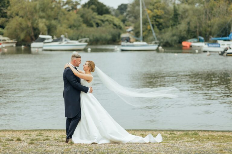 A bride and groom on the grass beside the River Stour, her veil lifting in the wind and sailing boats moored behind, Captain's Club Hotel & Spa