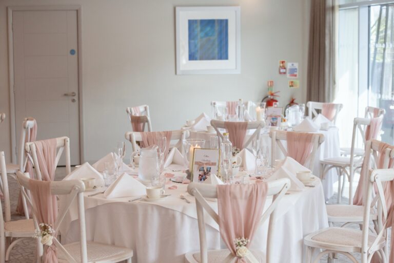 A function suite dressed for a wedding breakfast with blush chair sashes, white linen and small flower arrangements, Captain's Club Hotel & Spa